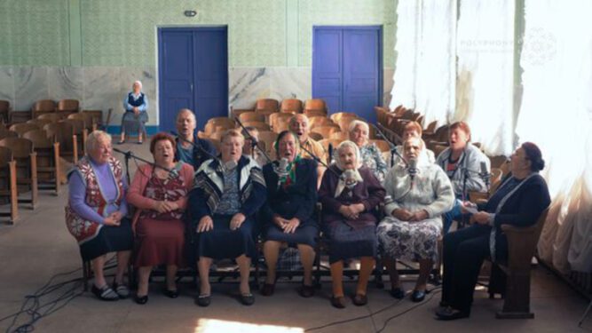 Ukranian women sitting and singing in front of microphones in a sunny room.